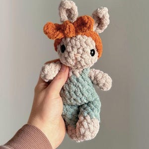 May include: A handmade crochet bunny doll with a light beige body, light blue overalls, and orange bow. The bunny has black eyes and pink accents on its feet. This soft toy is perfect for children.