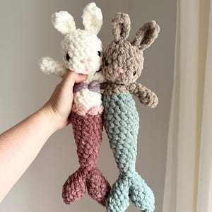 May include: Two crocheted mermaid bunny plushies. One is white with a pink tail and the other is brown with a teal tail.