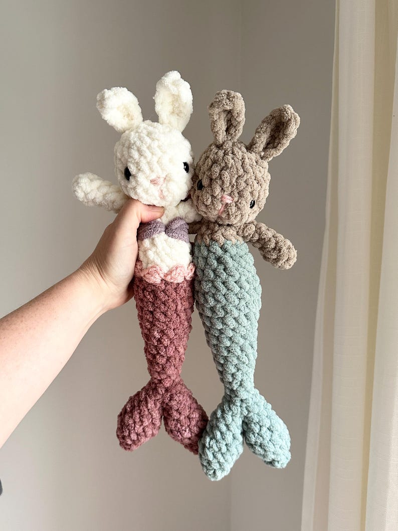 May include: Two crocheted mermaid bunny plushies. One is white with a pink tail and the other is brown with a teal tail.