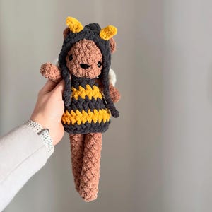 May include: A crocheted brown teddy bear wearing a black and yellow bee costume with a pointed hood and antennae.