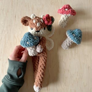 K&ouml;nnte beinhalten: Ein braunes und wei&szlig;es geh&auml;keltes Hirschspielzeug, das einen blauen und wei&szlig;en geh&auml;kelten Pilz h&auml;lt. Der Hirsch hat eine rosa Blume auf dem Kopf. Im Hintergrund befinden sich zwei weitere geh&auml;kelte Pilze, einer rosa und wei&szlig; und einer blau und wei&szlig;.