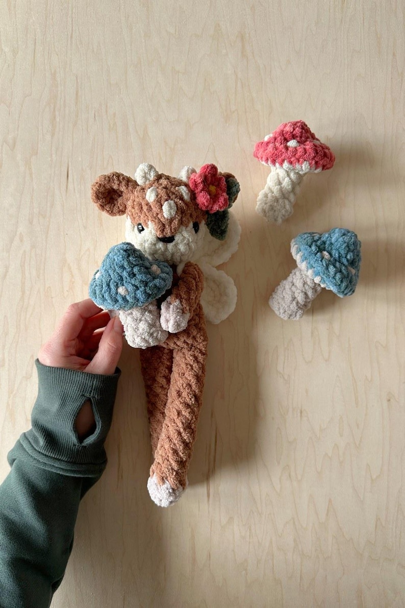 K&ouml;nnte beinhalten: Ein braunes und wei&szlig;es geh&auml;keltes Hirschspielzeug, das einen blauen und wei&szlig;en geh&auml;kelten Pilz h&auml;lt. Der Hirsch hat eine rosa Blume auf dem Kopf. Im Hintergrund befinden sich zwei weitere geh&auml;kelte Pilze, einer rosa und wei&szlig; und einer blau und wei&szlig;.