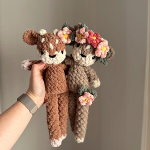 May include: Two crocheted deer plushies with floral crowns. The deer are brown and beige with white accents. The crowns are made of pink, red, and yellow flowers.