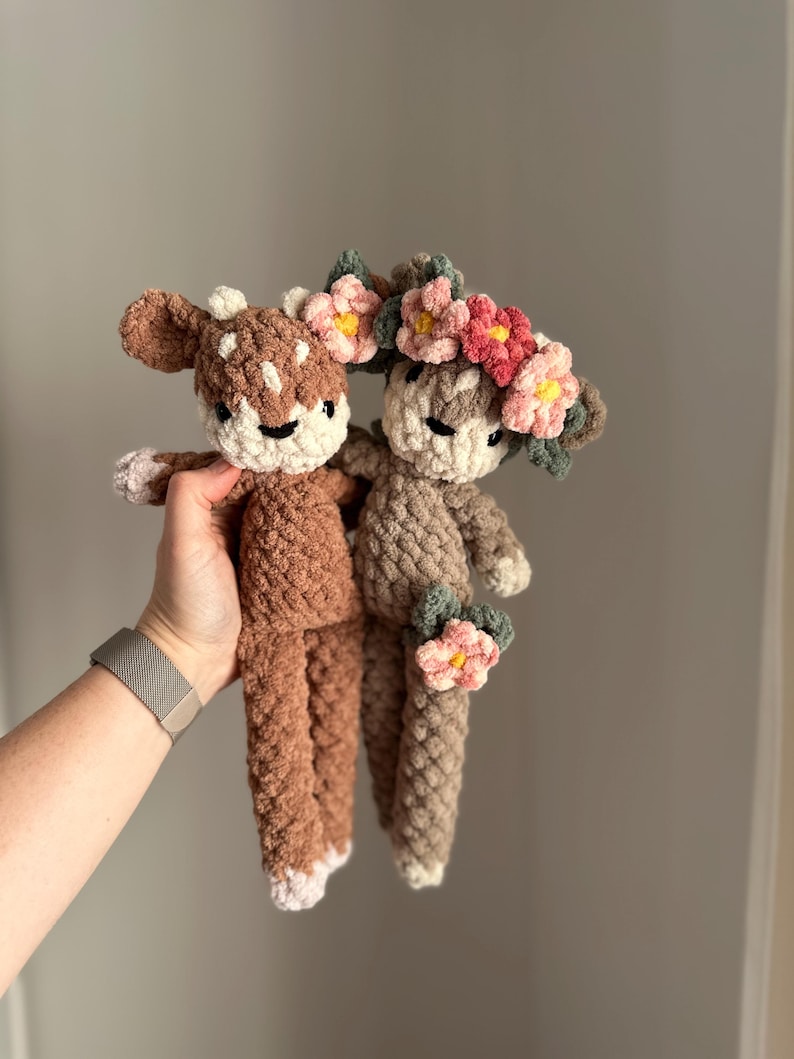 May include: Two crocheted deer plushies with floral crowns. The deer are brown and beige with white accents. The crowns are made of pink, red, and yellow flowers.