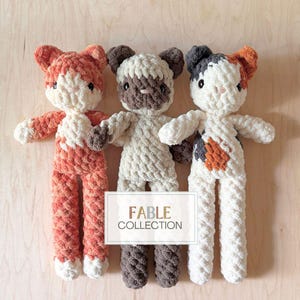 Op de afbeelding: Drie handgemaakte gehaakte kattenpoppen in oranje, crème en bruin. De poppen hebben slappe oren, armen en benen. De middelste pop is crème met bruine accenten, terwijl de andere twee oranje en grijze accenten hebben. De poppen zijn van de Fable Collection.