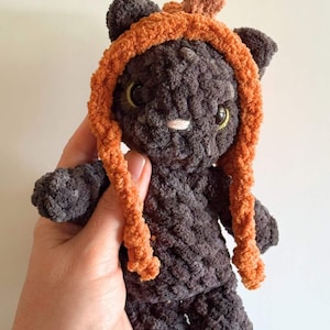 May include: A handmade, plush black cat doll with an orange hood and matching braids. The cat has yellow eyes and a white nose. The doll is made of a soft, textured material and is held in a person's hand.