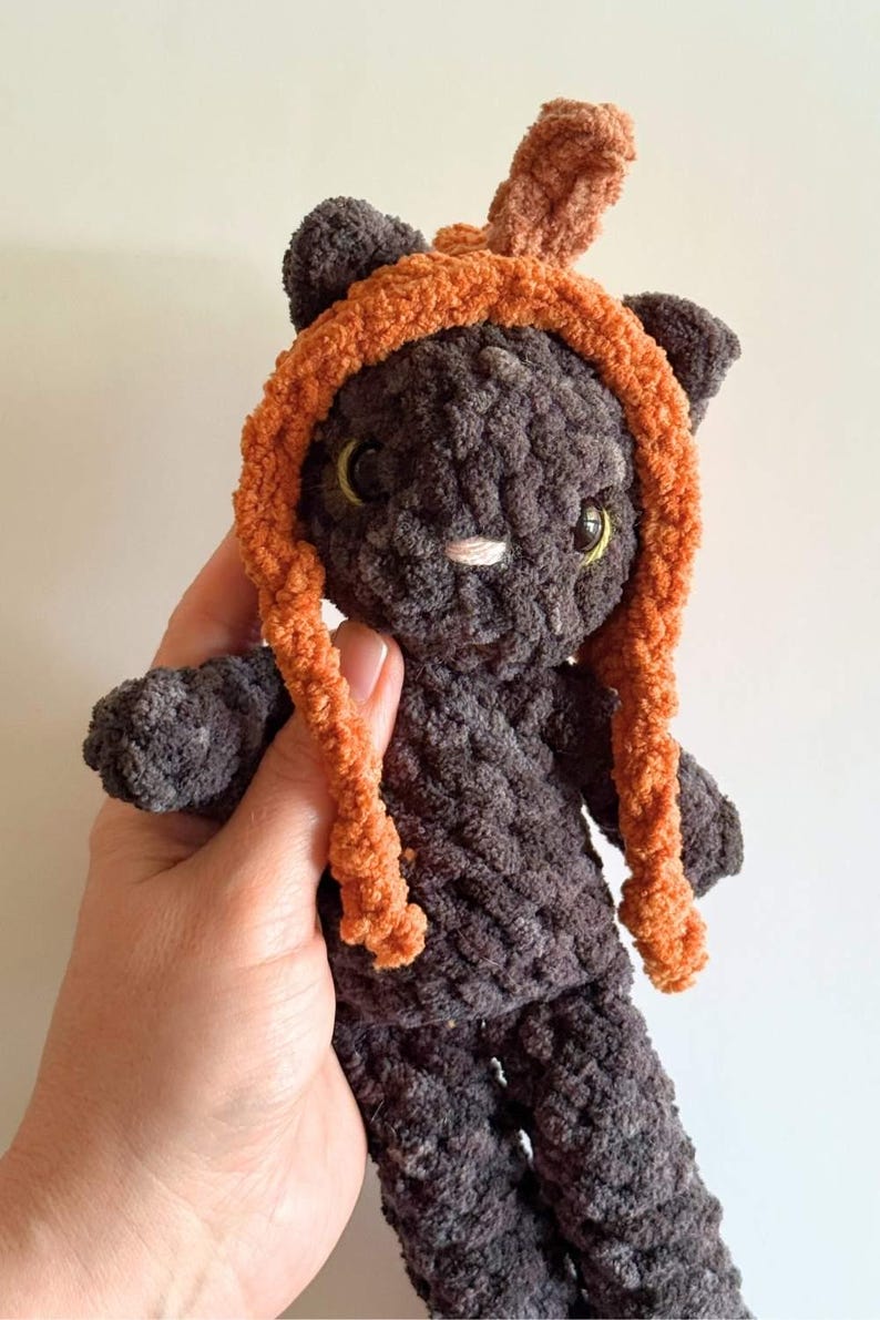 May include: A handmade, plush black cat doll with an orange hood and matching braids. The cat has yellow eyes and a white nose. The doll is made of a soft, textured material and is held in a person's hand.