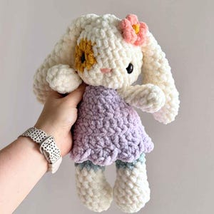 May include: A plush crochet bunny toy made of soft white yarn with long ears, a lavender dress, and a small pink flower accessory on its head. The bunny features a yellow patch over one eye, a small pink nose, and black bead eyes.