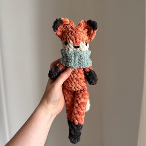May include: A crocheted fox plush toy with a light blue scarf. The fox is orange and black with a white belly.