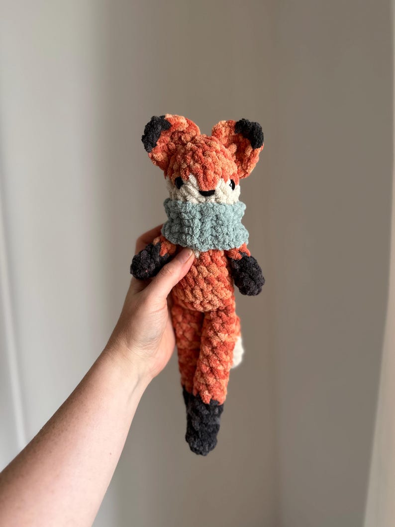 May include: A crocheted fox plush toy with a light blue scarf. The fox is orange and black with a white belly.