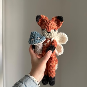 May include: A crocheted fox plush toy with orange and brown fur, black ears and nose, and white wings. The fox is holding a blue and green mushroom.