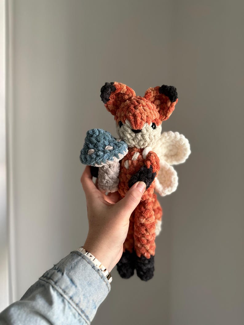 May include: A crocheted fox plush toy with orange and brown fur, black ears and nose, and white wings. The fox is holding a blue and green mushroom.