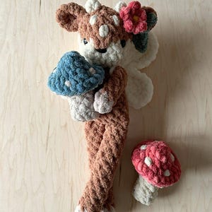 K&ouml;nnte beinhalten: Ein geh&auml;keltes Pl&uuml;schtier in Form eines Rehs mit einer Blume auf dem Kopf, das einen blauen Pilz h&auml;lt. Ein rosa Pilz liegt auf der Holzoberfl&auml;che.