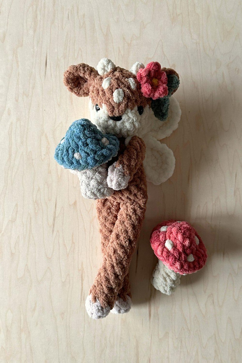 K&ouml;nnte beinhalten: Ein geh&auml;keltes Pl&uuml;schtier in Form eines Rehs mit einer Blume auf dem Kopf, das einen blauen Pilz h&auml;lt. Ein rosa Pilz liegt auf der Holzoberfl&auml;che.