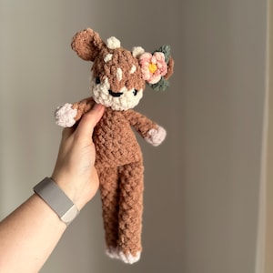 May include: A brown crocheted deer plush toy with a pink flower and green leaves on its head. The deer has white accents on its face and feet.