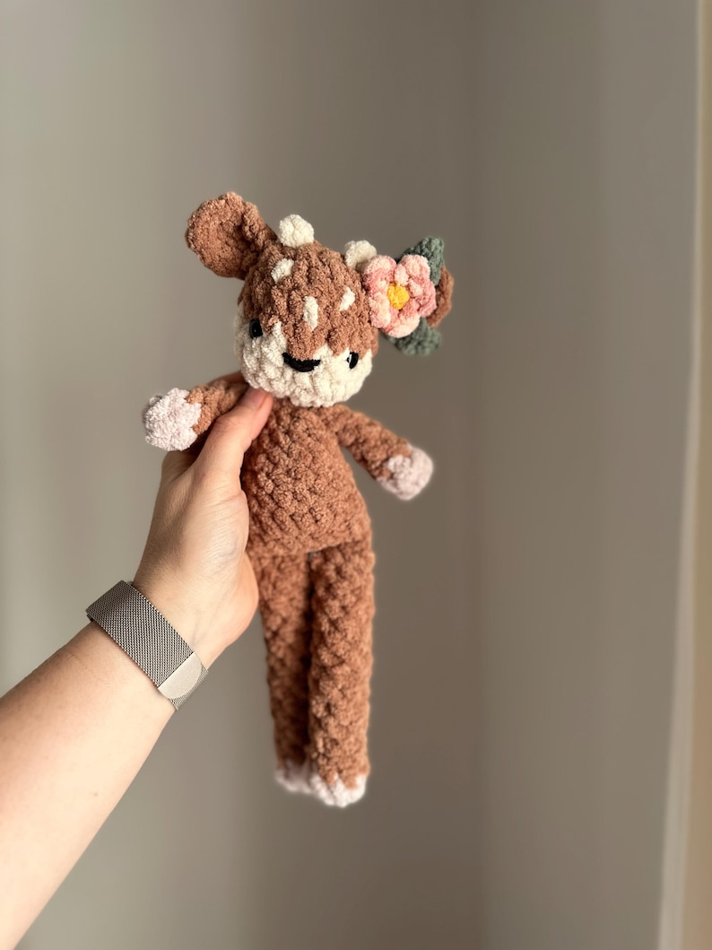 May include: A brown crocheted deer plush toy with a pink flower and green leaves on its head. The deer has white accents on its face and feet.