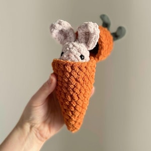 May include: A crocheted carrot with a small pink bunny peeking out of the top. The carrot is orange and the bunny has black eyes.