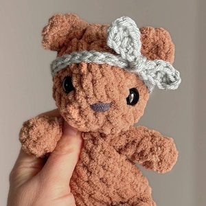 May include: A handmade, brown crochet teddy bear with a gray headband. The bear has black safety eyes and a purple stitched mouth. The soft, textured yarn gives the bear a cuddly appearance. A charming, handcrafted plush toy.