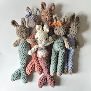 May include: A group of six crocheted plush bunny toys. The bunnies are in various colours, including brown, grey, white, orange, and blue. Some of the bunnies have mermaid tails.