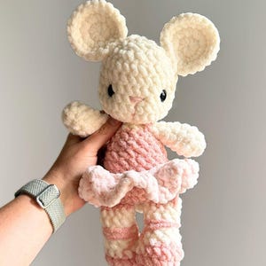 May include: A plush crochet mouse toy featuring a cream-colored head with large round ears and black eyes. The body is dressed in a soft pink crocheted tutu and matching pink leg warmers on the feet, crafted from a thick, textured yarn material.