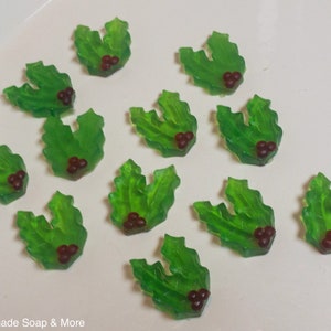 May include: Twelve green holly leaf-shaped soaps with red berries. The soaps are arranged on a white surface.