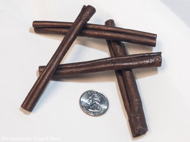 May include: Four brown cinnamon sticks arranged in a cross pattern on a white surface. A US quarter is placed in the center for size comparison.