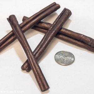 May include: Five brown cinnamon sticks arranged in a criss-cross pattern on a white surface. A US quarter is included for scale.