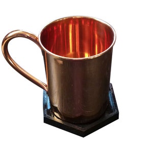 May include: A copper mug with a handle sits on a black hexagonal coaster. The mug is empty and has a shiny, reflective surface.