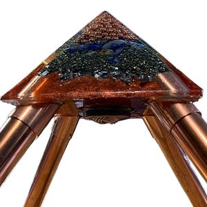 May include: A pyramid-shaped object featuring a clear resin top embedded with small copper spheres, blue lapis lazuli stones, and dark metallic granules. The base is supported by four polished copper tubes that extend outward from the center.