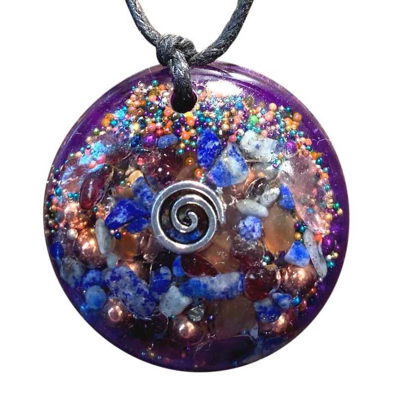May include: A round, purple, resin pendant with a silver spiral charm. The pendant is filled with various crystals, including blue lapis lazuli, copper, and clear quartz.
