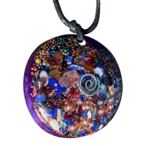 May include: A round, purple, resin pendant with a silver spiral charm. The pendant is filled with various crystals, including blue, red, and gold.