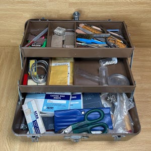 May include: A brown metal cantilever tool box with three tiers containing various jewelry making supplies. The compartments hold items including Kerr casting wax sheets, metal shears, wire, files, small containers, and assorted hand tools.