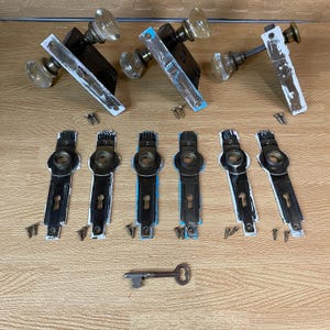 May include: A collection of vintage door hardware, including glass doorknobs, black and white painted door plates, and a skeleton key. The hardware is arranged on a light wood surface, with screws scattered around the pieces.
