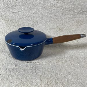 May include: A deep blue enameled saucepan with a matching lid and a long wooden handle. The pot features a small pouring spout on the side and a circular knob on the lid. The exterior finish is glossy, and the handle has a metallic end cap.