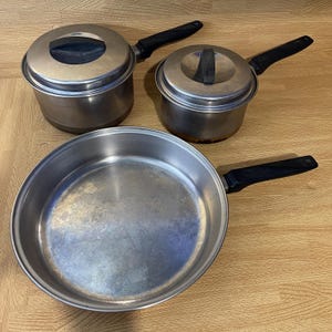 May include: A set of three stainless steel cookware pieces including two lidded saucepans and one large frying pan. Each item features a polished silver finish and a black heat-resistant handle. The lids have black knobs and fit securely.