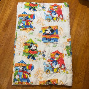 Puede incluir: Manta acolchada blanca con un estampado colorido con tem&aacute;tica de carnaval con Mickey Mouse, Donald Duck y Goofy. El dise&ntilde;o incluye carpas de circo, globos y puestos de dulces. La manta es rectangular.