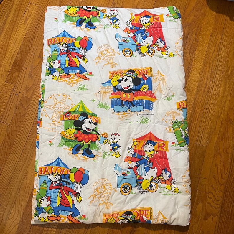 Puede incluir: Manta acolchada blanca con un estampado colorido con tem&aacute;tica de carnaval con Mickey Mouse, Donald Duck y Goofy. El dise&ntilde;o incluye carpas de circo, globos y puestos de dulces. La manta es rectangular.