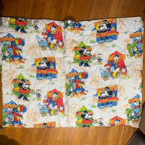 Puede incluir: S&aacute;bana blanca con un estampado repetido de Mickey Mouse, el Pato Donald y otros personajes de Disney en un carnaval. Las ilustraciones incluyen tiendas de campa&ntilde;a, globos y carritos de caramelos en colores brillantes como el rojo, el azul y el amarillo. La s&aacute;bana est&aacute; arrugada.