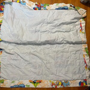 Puede incluir: Una manta acolchada azul claro con un borde blanco con personajes de dibujos animados y veh&iacute;culos de varios colores. La manta est&aacute; doblada por la mitad, revelando el interior acolchado azul claro.