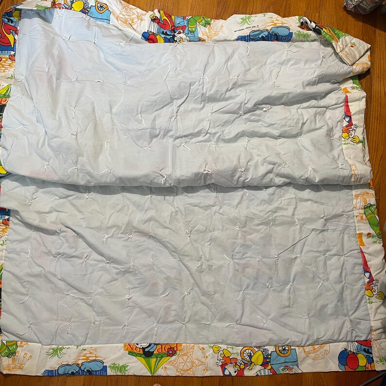 Puede incluir: Una manta acolchada azul claro con un borde blanco con personajes de dibujos animados y veh&iacute;culos de varios colores. La manta est&aacute; doblada por la mitad, revelando el interior acolchado azul claro.