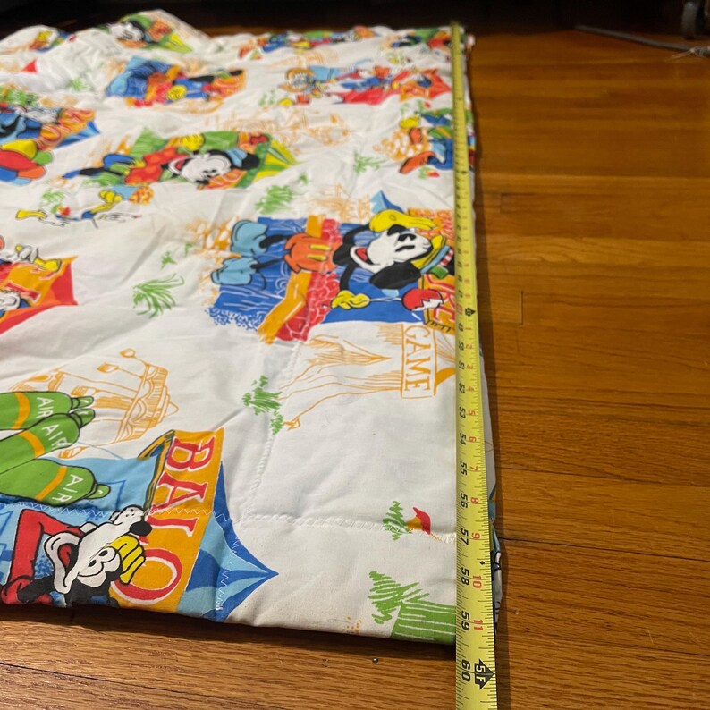 Puede incluir: Tela blanca con un estampado colorido con Mickey Mouse y otros personajes de Disney. La tela se mide con una cinta m&eacute;trica amarilla, mostrando una longitud de aproximadamente 152 cm.