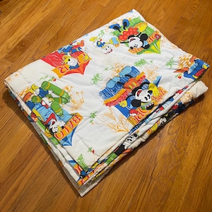 Puede incluir: Un edred&oacute;n blanco plegado con personajes de dibujos animados coloridos, incluyendo Mickey Mouse y el Pato Donald, en varias escenas l&uacute;dicas. El edred&oacute;n tiene un patr&oacute;n repetitivo con las palabras "OPEN GAME" y "BALO".