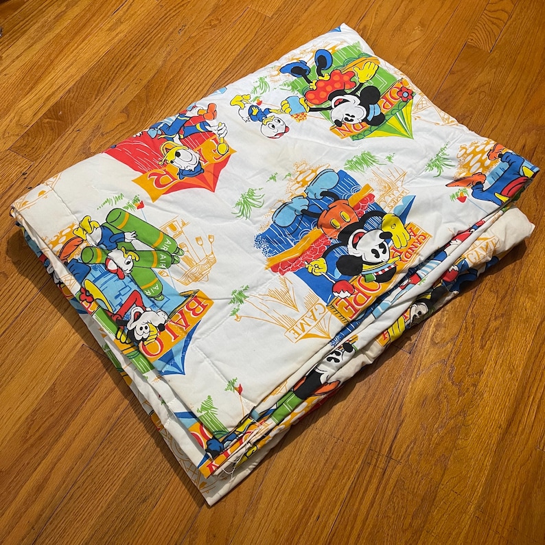 Puede incluir: Un edred&oacute;n blanco plegado con personajes de dibujos animados coloridos, incluyendo Mickey Mouse y el Pato Donald, en varias escenas l&uacute;dicas. El edred&oacute;n tiene un patr&oacute;n repetitivo con las palabras "OPEN GAME" y "BALO".