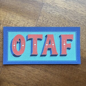 May include: A rectangular sticker with the letters "OTAF" in red, outlined in white, on a light blue background. The letters have a dotted pattern. The background is framed by a dark blue border.