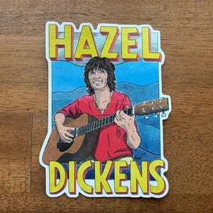 Puede incluir: Una pegatina con una colorida ilustración de Hazel Dickens tocando la guitarra. La pegatina tiene el texto "HAZEL" en letras amarillas en bloque encima de la imagen y "DICKENS" debajo. El fondo es azul y la pegatina está delineada en blanco.