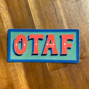 May include: A blue and green rectangular sign with the word "OTAF" in red letters with white dots.