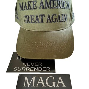 May include: A green baseball cap with the text "MAKE AMERICA GREAT AGAIN" embroidered in black. The cap is sitting on top of two black stickers with the text "Maga Never Surrender" in white.
