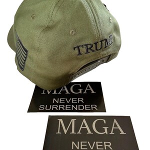 May include: A green baseball cap with a black embroidered American flag on the side and the word "TRUMP" embroidered on the back. Two black stickers with white text that reads "MAGA NEVER SURRENDER" are included.