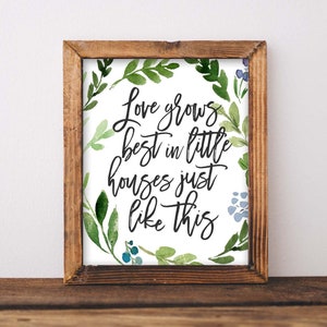 May include: A framed print with a white background and a green watercolour wreath. The wreath has blue flowers and green leaves. The text in the centre reads "Love grows best in little houses just like this."