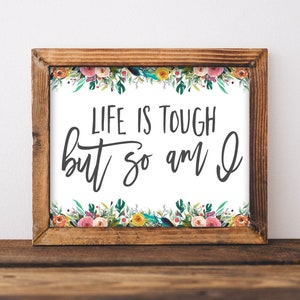 May include: A framed print with a white background and a floral border. The text "Life is tough but so am I" is written in black cursive font.